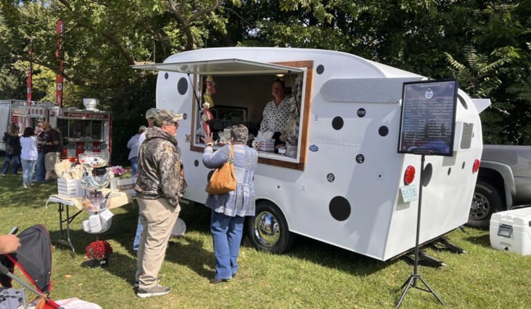 White polka dotted food trailer serving customers at an outdoor park with a menu board and people in line