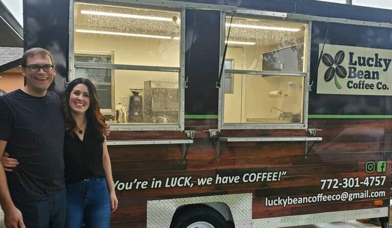 Two people smiling beside a wooden coffee truck with the Lucky Bean Coffee Co logo and slogan visible in the side panel