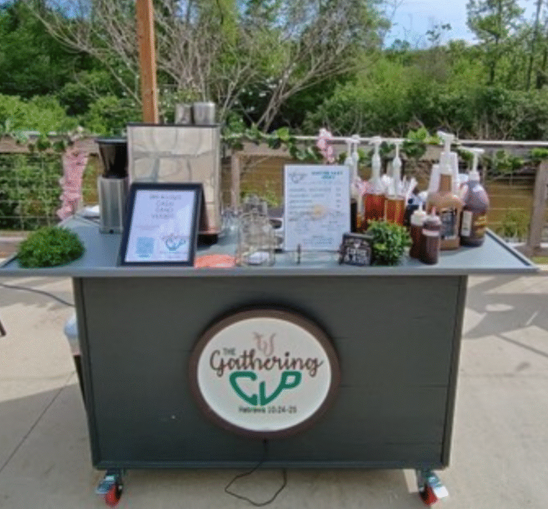 Outdoor mobile drink cart branded The Gathering on a patio with syrups bottles signs and a small plant nearby